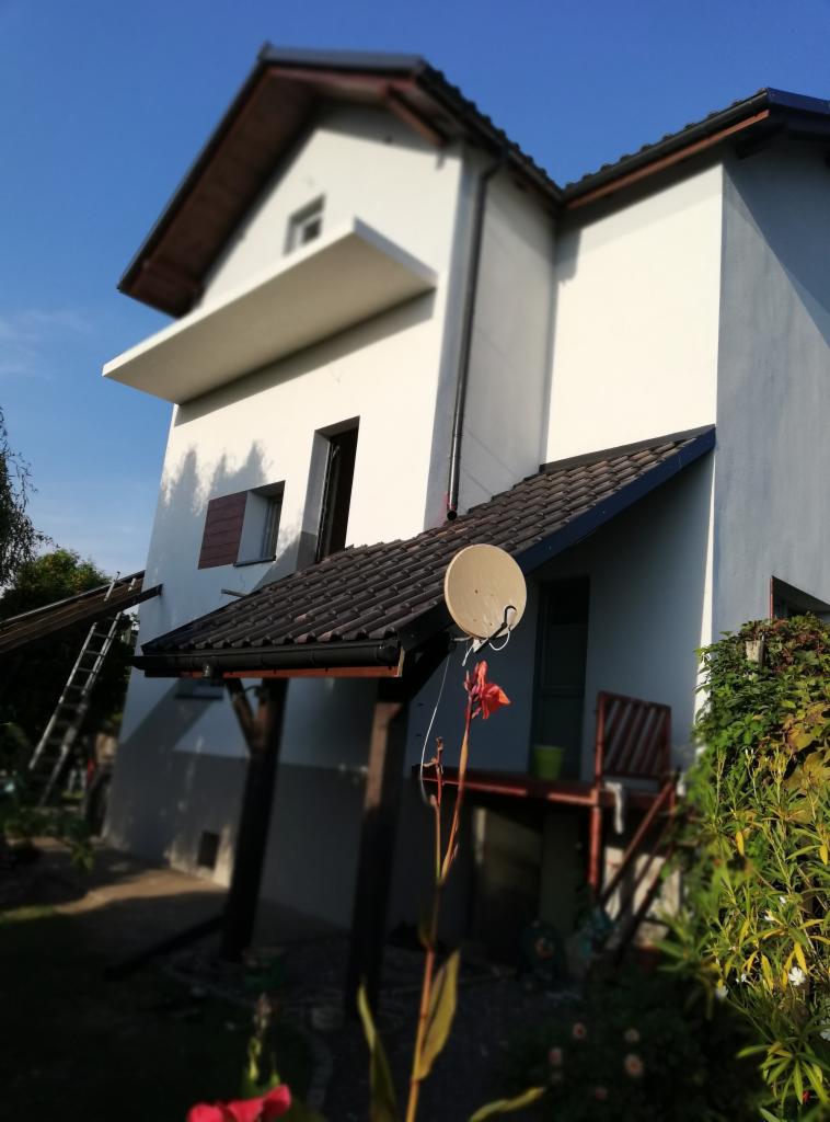 Dwupiętrowy dom z nową, białą elewacją, widoczny z boku, z anteną satelitarną na dachu ganku i drabiną opartą o ścianę, sugerującą trwające prace.