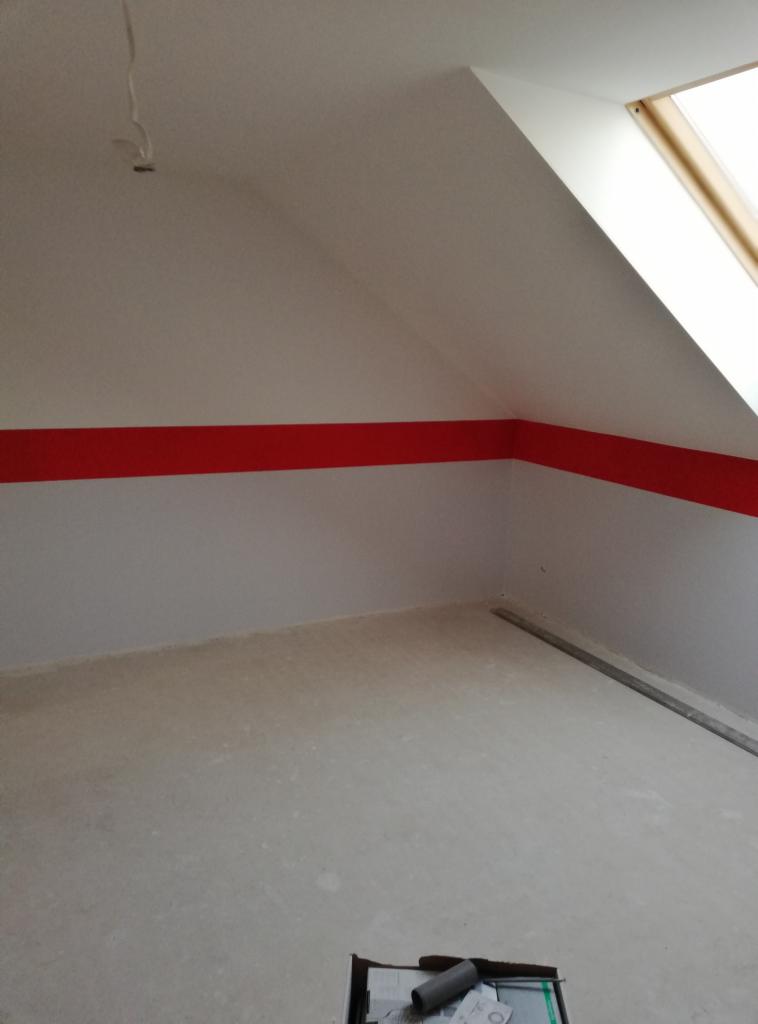 Poddasze po malowaniu, białe ściany z poziomym czerwonym pasem, okno dachowe, surowa podłoga przed wykończeniem.