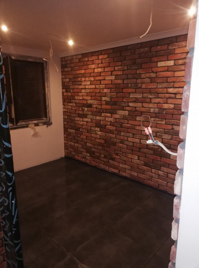 Pokój po remoncie z imitacją ceglanej ściany, ciemną podłogą z płytek i widocznymi przewodami elektrycznymi.