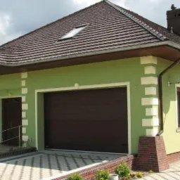 Zakończona budowa domu jednorodzinnego z brązowym dachem z dachówki, zieloną elewacją z dekoracyjnymi białymi elementami, brązową bramą garażową i satelitą na ścianie.