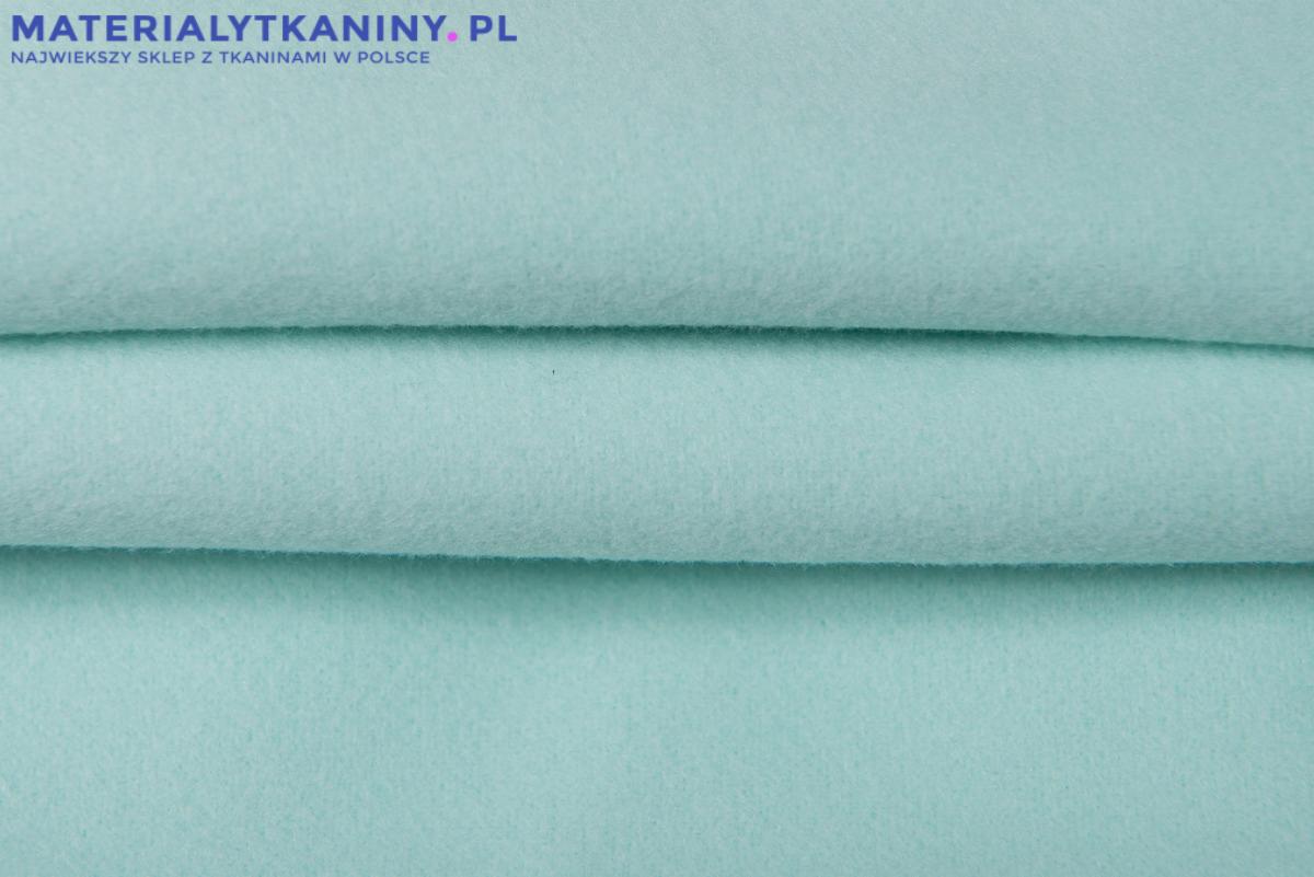 Jasnoturkusowa, miękka tkanina polarowa, ułożona w fałdy, z widocznym logo sklepu internetowego w górnym lewym rogu.