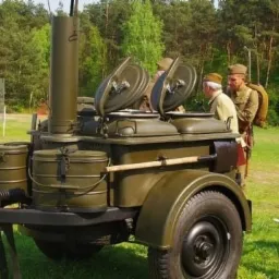 Polowa kuchnia wojskowa z otwartymi pokrywami, przygotowana do wydawania posiłków, w tle żołnierze i rower z flagą Polski.