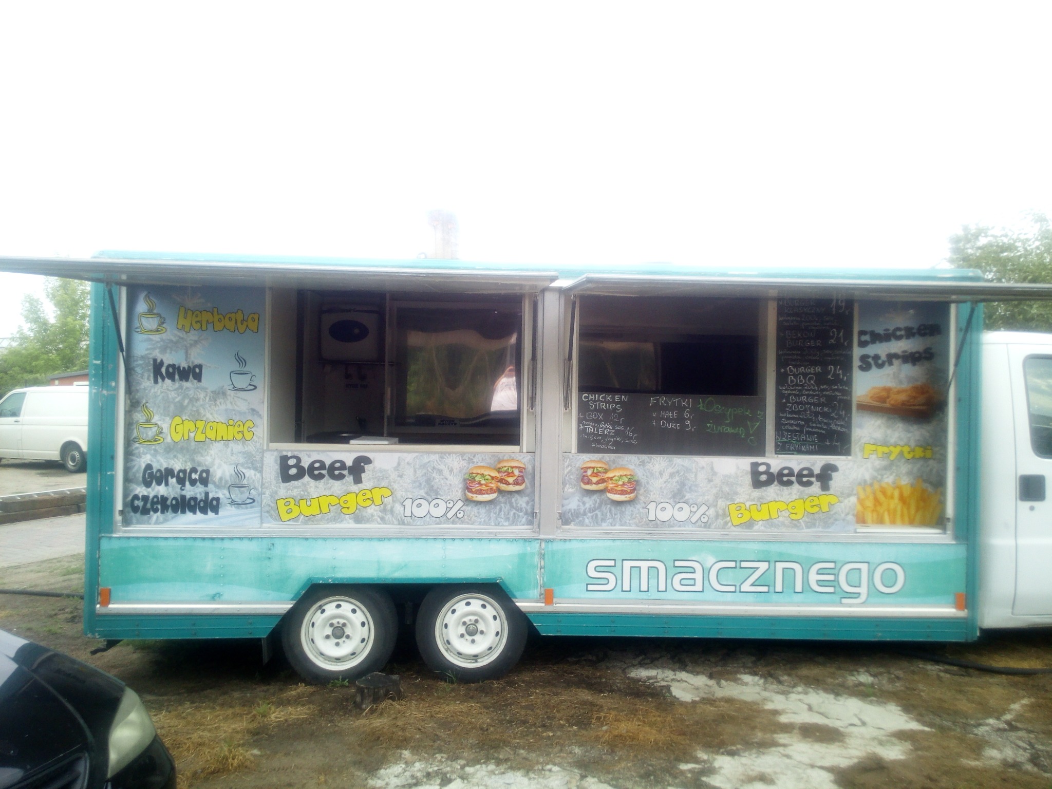 Turkusowa przyczepa gastronomiczna z menu wypisanym na tablicy kredowej i naklejkami burgerów, oferująca herbatę, kawę, grzańca, czekoladę, burgery wołowe i frytki.