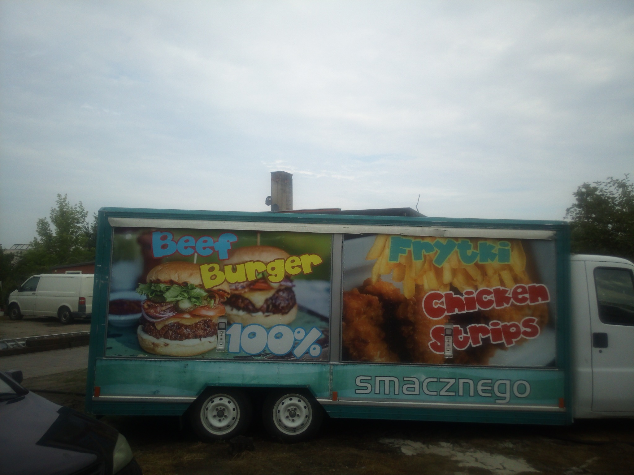 Przyczepa gastronomiczna z reklamą burgerów i frytek z kurczakiem, zaparkowana na zewnątrz, widoczny napis 'smacznego'.