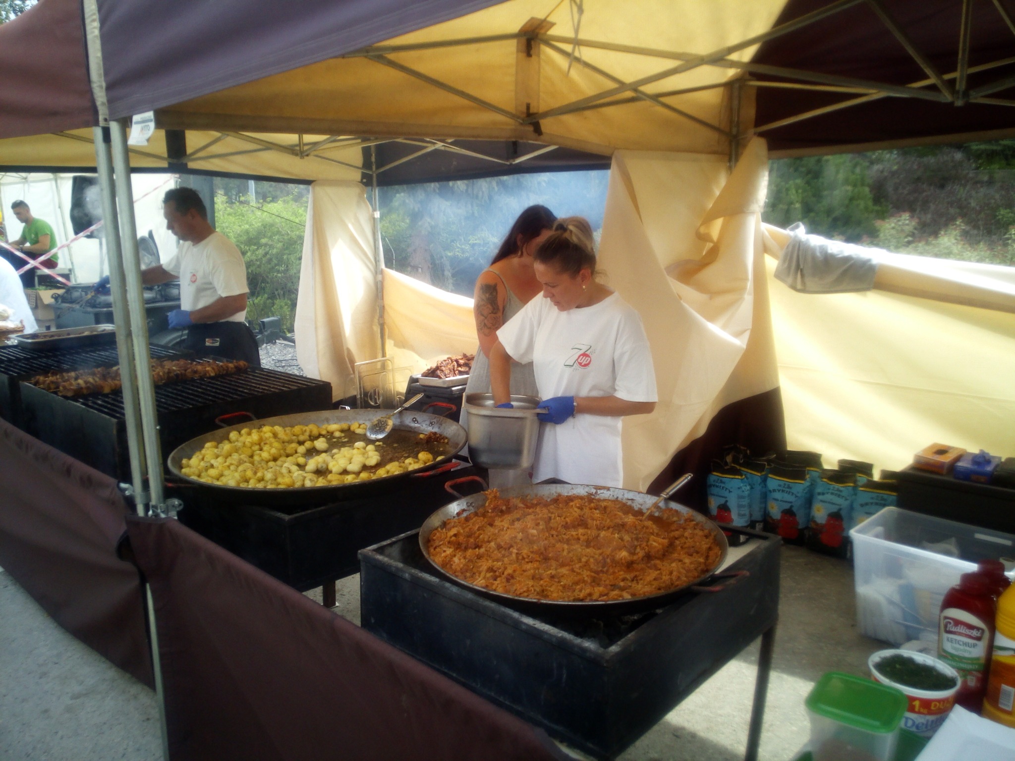 Przygotowywanie potraw na dużych patelniach pod zadaszeniem, widoczne ziemniaki, bigos, grillowane szaszłyki, pracownicy w białych koszulkach i niebieskich rękawiczkach.