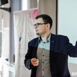 Prelegent w okularach, w trakcie prezentacji, wskazujący palcem do góry na ekran z napisem 'uwagę na włas'