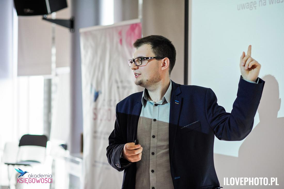 Prelegent w okularach, w trakcie prezentacji, wskazujący palcem do góry na ekran z napisem 'uwagę na włas'