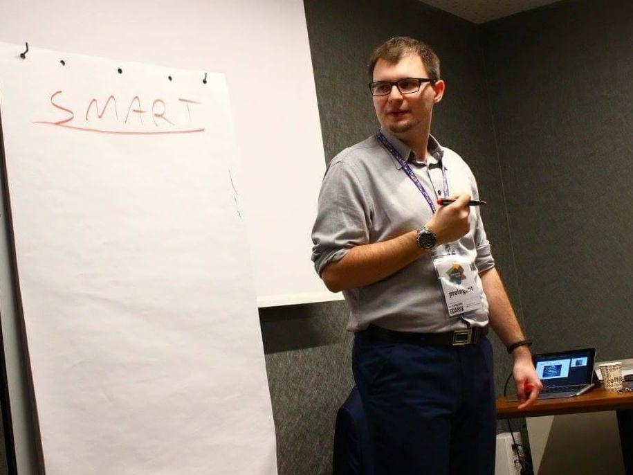 Prelegent z markerem w dłoni stoi obok flipcharta z napisem 'SMART' podczas prezentacji. Na stole laptop i kubek z kawą.