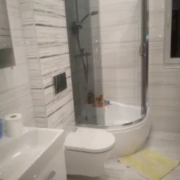 Wykończona łazienka z narożną kabiną prysznicową, wiszącą toaletą i umywalką z szafką, wyłożona płytkami imitującymi biały marmur z szarymi żyłami, z żółtą wycieraczką na podłodze.