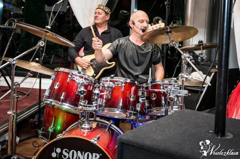Zespół muzyczny podczas występu: perkusista z mikrofonem i gitarzysta w czarnym stroju i ozdobnym nakryciu głowy, czerwona perkusja marki Sonor.
