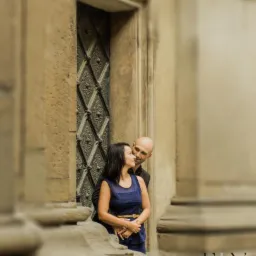 Para zakochanych stoi w portalu ozdobnych drzwi, kobieta w niebieskiej sukience z paskiem, mężczyzna łysy w ciemnej marynarce, dekoracyjny portal, oświetlenie naturalne.