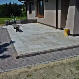 Wyłożony jasnoszarą kostką brukową taras przydomowy z obrzeżem z ciemniejszej kostki, widoczny fragment ścieżki z kostki oraz żółty stołek obok kolumny domu w Katowicach.