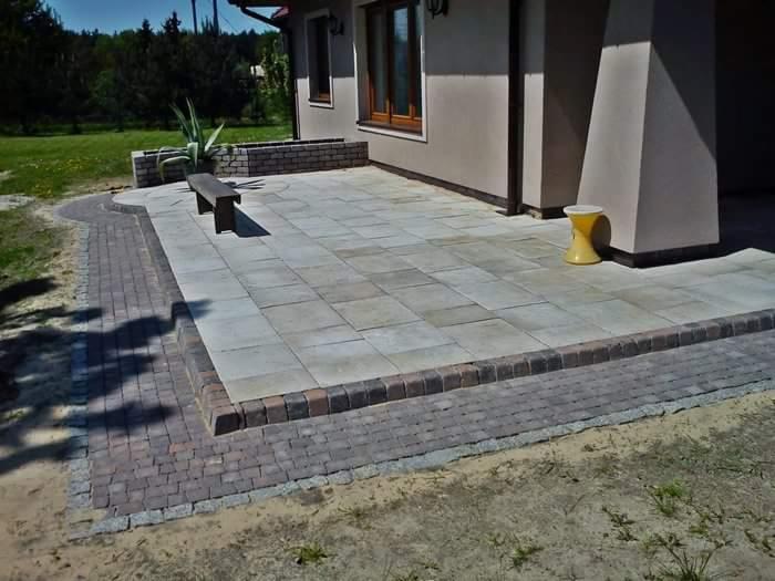 Wyłożony jasnoszarą kostką brukową taras przydomowy z obrzeżem z ciemniejszej kostki, widoczny fragment ścieżki z kostki oraz żółty stołek obok kolumny domu w Katowicach.