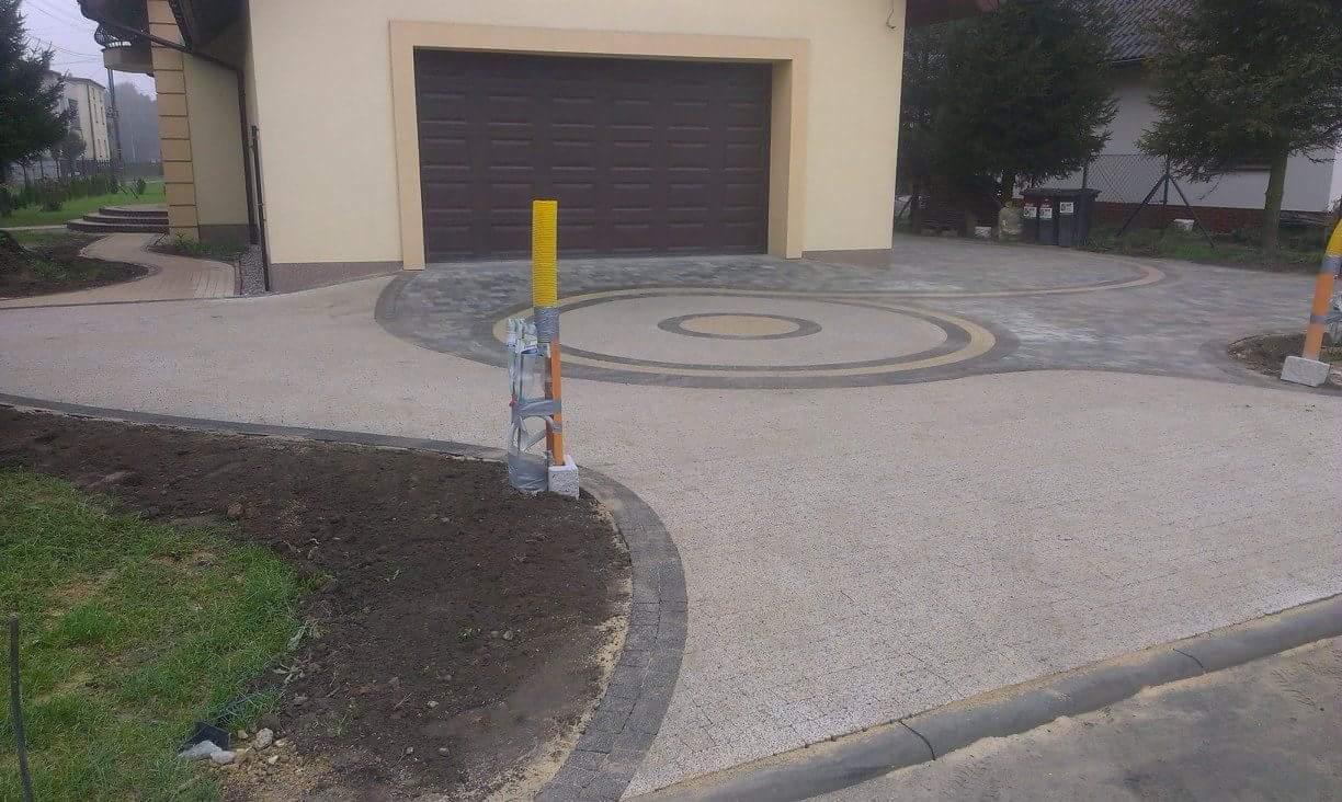 Nowo ułożony podjazd z jasnej kostki brukowej z ciemnymi obramowaniami i okrągłym wzorem przed garażem, widoczne słupki zabezpieczające.