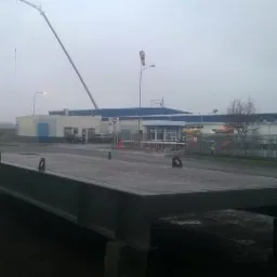 Metalowa platforma na betonowych podporach z uchwytami, przygotowana do transportu lub montażu, na tle przemysłowego krajobrazu z budynkami, dźwigiem i ciężarówką w pochmurny dzień.