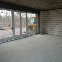 Wnętrze surowego domu w trakcie budowy z zamontowanymi czteroskrzydłowymi oknami z białymi ramami, widok na plac budowy z koparką i stertą desek.