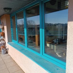 Okna-Plast Dariusz Wroński - Turkusowe okna z widokiem na balkon i miasto w tle. Na balkonie widoczny regał z doniczkami i konewka. Jasne, naturalne światło.