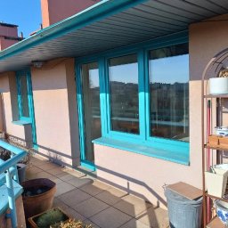 Okna-Plast Dariusz Wroński - Balkon z turkusowymi oknami i balustradą, widok na miasto w tle. Na balkonie donice z roślinami i metalowy regał z dekoracjami.