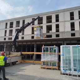 OKNA-PLAST - Okna, montaż i serwis Warszawa - Dźwig budowlany podnosi pakiet nowych, zafoliowanych okien na palecie, przygotowanych do montażu w otworach wielopiętrowego budynku mieszkalnego z białych pustaków. Na pierwszym planie pracownik...