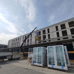 OKNA-PLAST - Okna, montaż i serwis Warszawa - Dźwig unosi dużą, białą ramę okienną w kierunku piętra niedokończonego budynku. Na pierwszym planie stosy zafoliowanych, nowych ram okiennych na paletach.