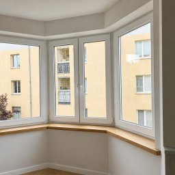 OKNA-PLAST - Okna, montaż i serwis Warszawa - Nowoczesne, białe okna w wykuszu z jasnymi, drewnianymi parapetami, z widokiem na żółty budynek mieszkalny.