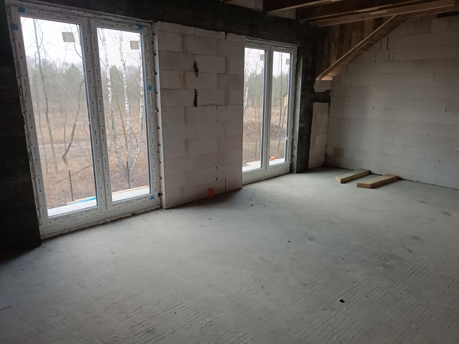 Surowe wnętrze w trakcie budowy z trzema białymi oknami z widokiem na las, ściany z białego bloczka, betonowa podłoga i drewniane belki stropowe.
