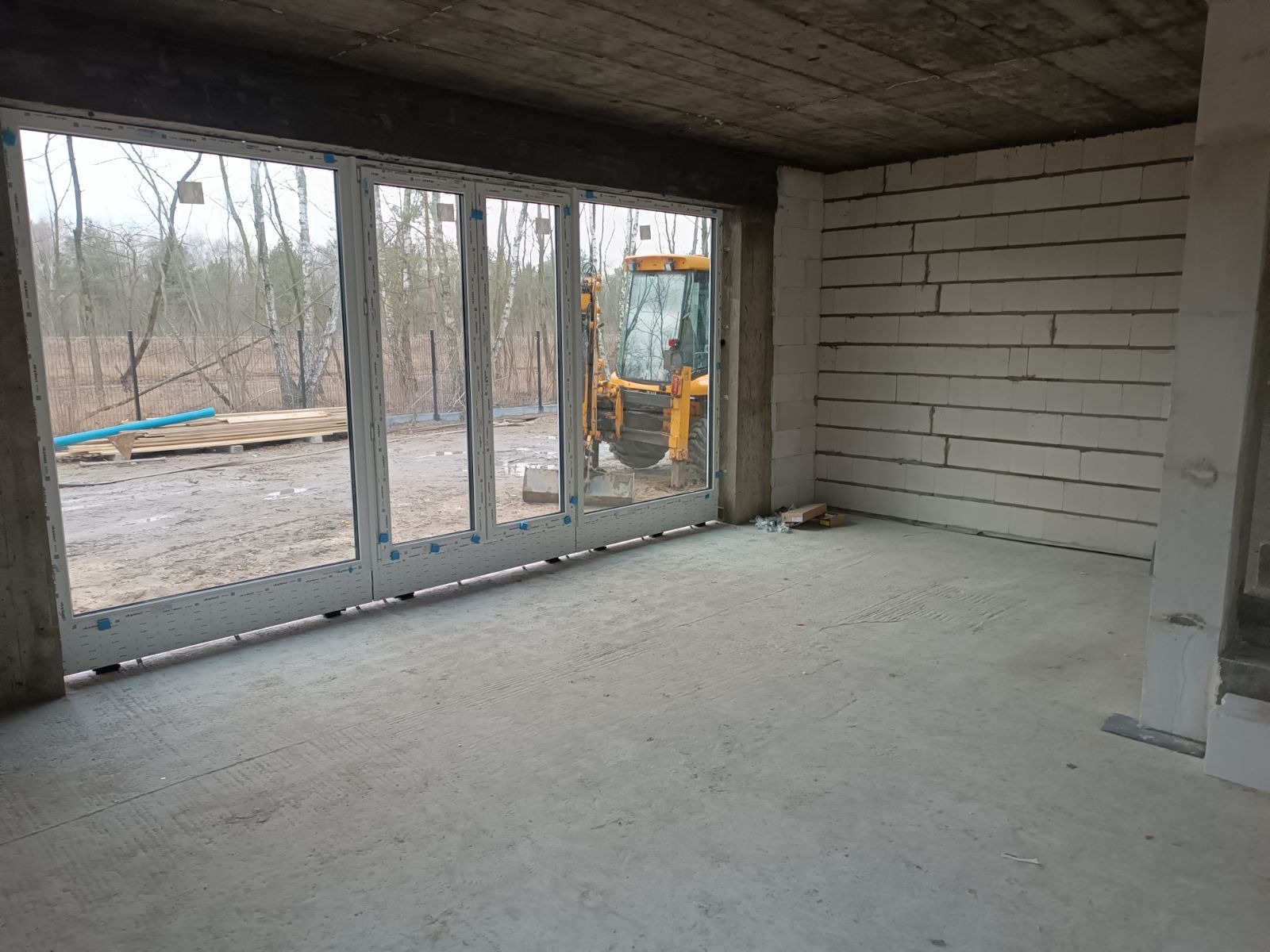 Wnętrze surowego domu w trakcie budowy z zamontowanymi czteroskrzydłowymi oknami z białymi ramami, widok na plac budowy z koparką i stertą desek.