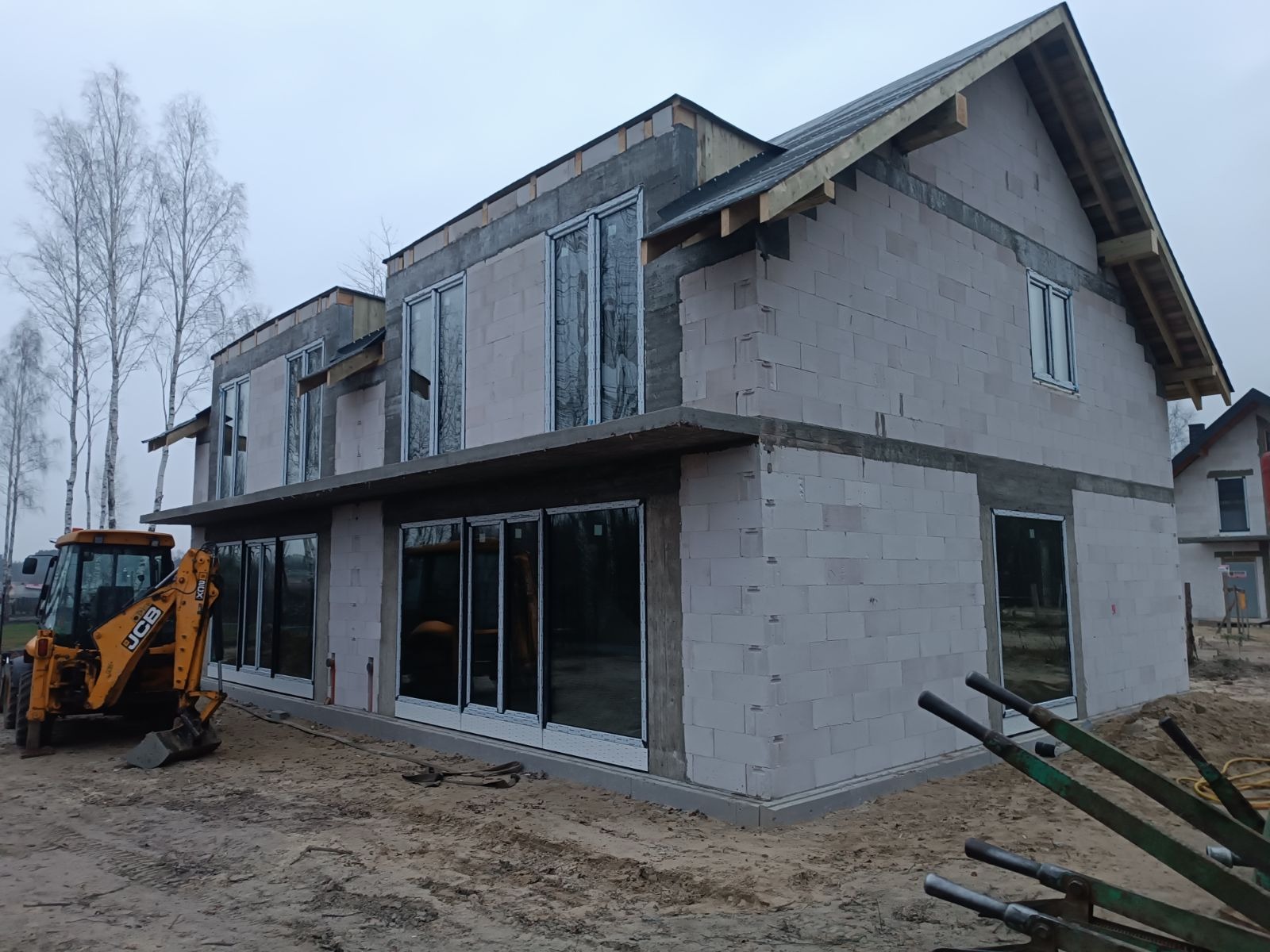 Budowa domu w Warszawie z zamontowanymi oknami PCV i aluminiowymi. Widoczny surowy stan budynku z bloczków, okna zabezpieczone folią, na pierwszym planie koparka JCB.