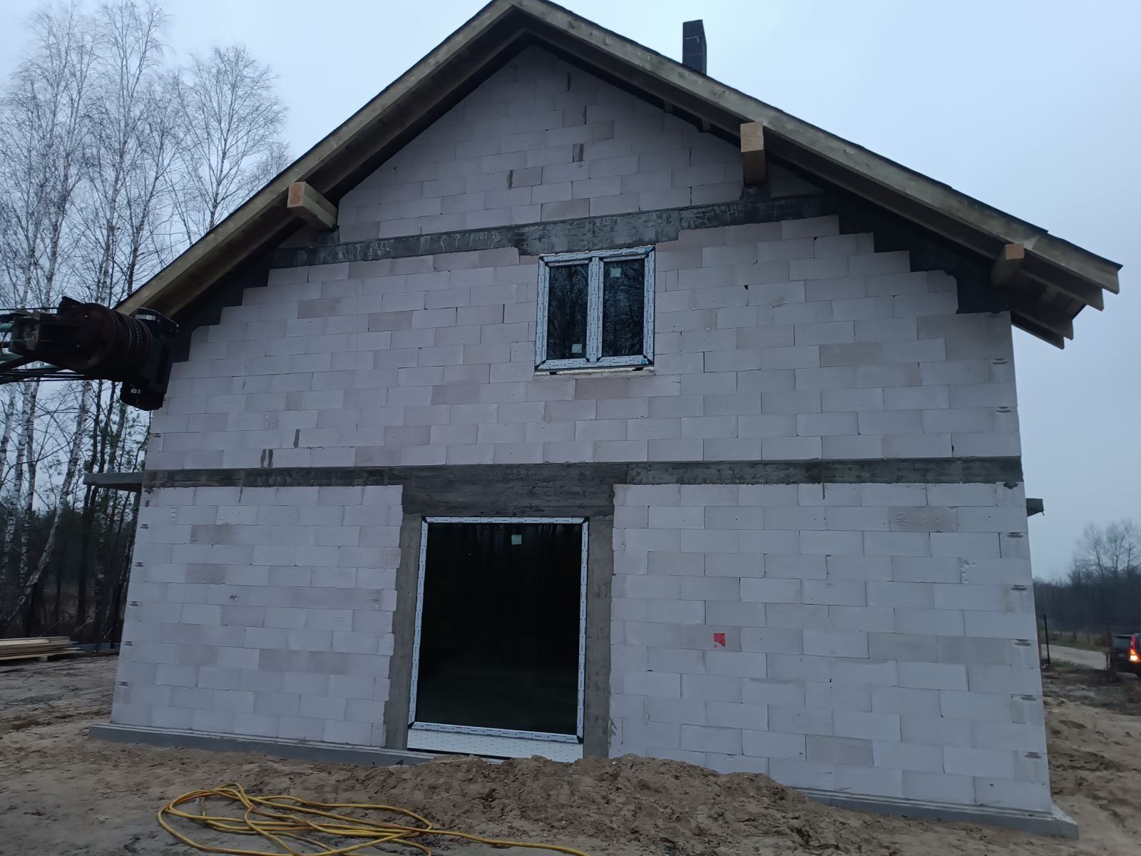 Surowy stan budynku w trakcie budowy z zamontowanymi oknami i przygotowanym otworem na drzwi, ściany z białych bloczków, widoczne elementy konstrukcji dachu i sprzęt budowlany.