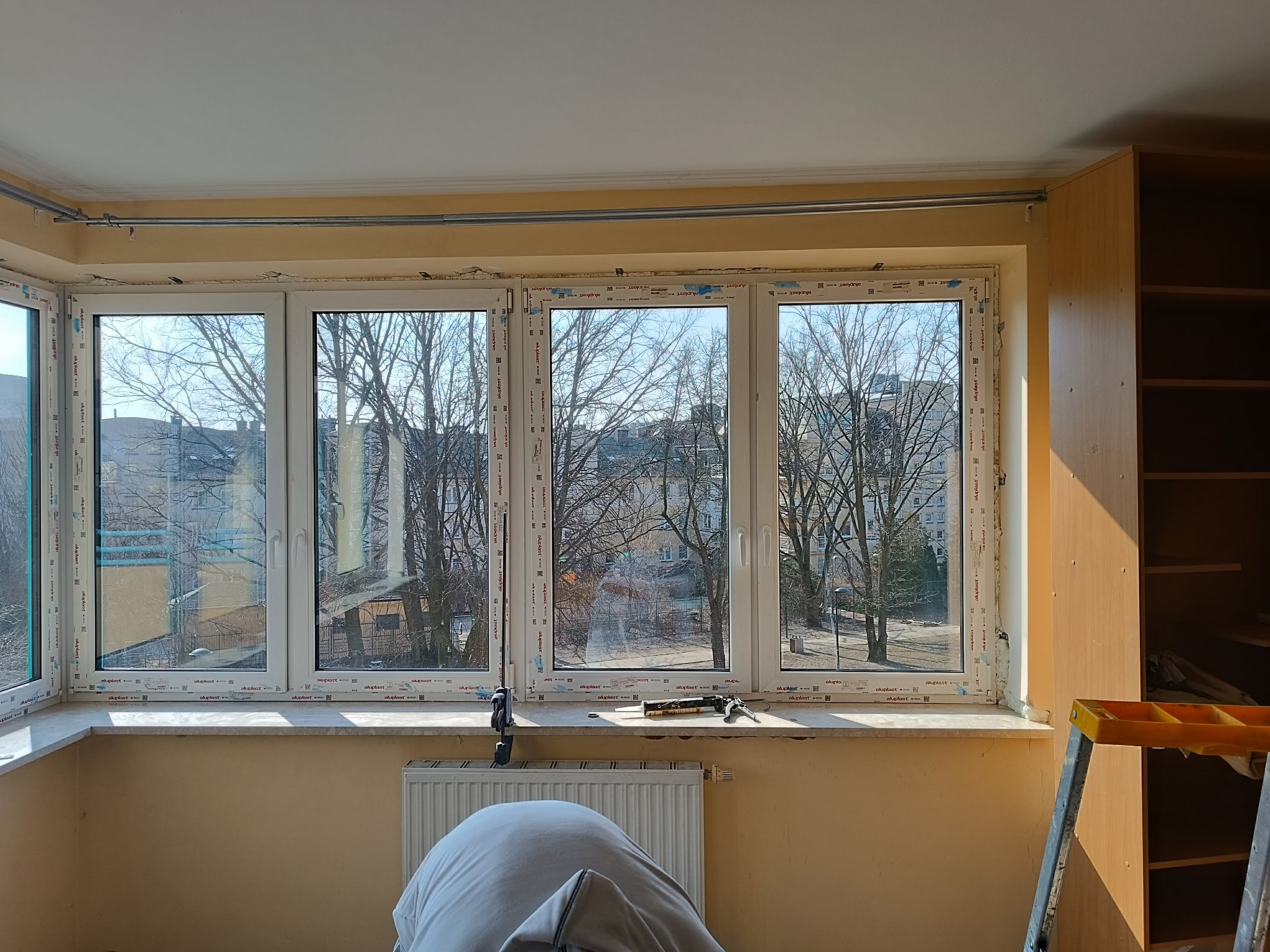 Jasne wnętrze z oknem PCV po montażu, widok na drzewa. Na parapecie narzędzia, pod oknem grzejnik. Ślady pianki montażowej wokół ramy okiennej.