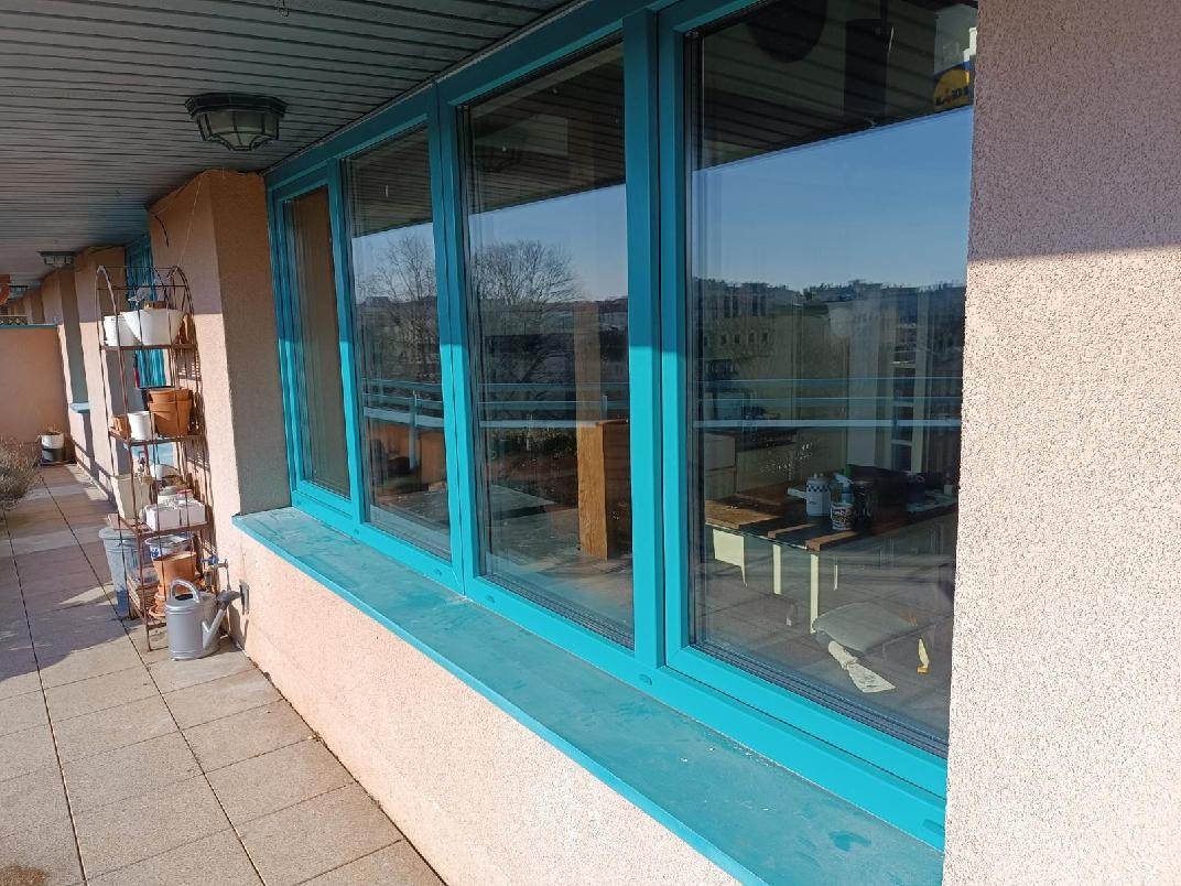 Turkusowe okna z widokiem na balkon i miasto w tle. Na balkonie widoczny regał z doniczkami i konewka. Jasne, naturalne światło.