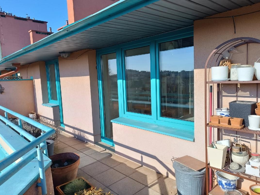 Balkon z turkusowymi oknami i balustradą, widok na miasto w tle. Na balkonie donice z roślinami i metalowy regał z dekoracjami.