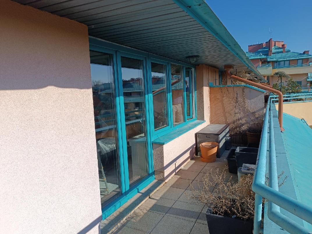 Balkon z turkusowymi oknami PCV i balustradą, widok na budynek w tle. Jasne, słoneczne oświetlenie. Widoczne donice i elementy wyposażenia balkonu.