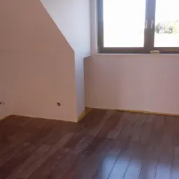 Poddasze z ukończonymi panelami podłogowymi w odcieniach brązu, świeżo pomalowane ściany na biało z widocznymi otworami na gniazdka elektryczne, okno z widokiem na zieleń.