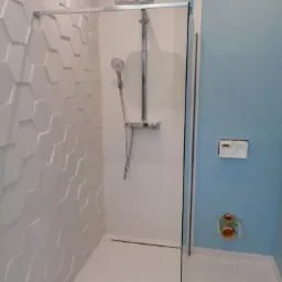 Nowoczesna kabina prysznicowa z geometryczną białą ścianą i błękitną ścianą z widocznymi przyłączami hydraulicznymi.