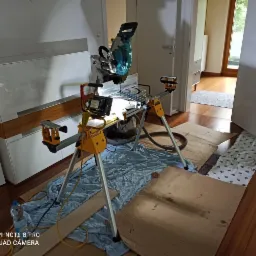 Piła ukośnica DeWalt na statywie w trakcie pracy, zabezpieczone podłoże, widoczne elementy podłogowe w pomieszczeniu z drewnianą podłogą i białymi meblami.