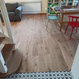 Jasne panele podłogowe ułożone w salonie, widoczne przejście między panelami a wzorzystą, czarno-białą podłogą z płytek, fragment schodów z białymi balustradami po lewej stronie.