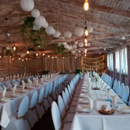 Dekoracja sali weselnej w stylu rustykalnym: długie stoły z białymi obrusami, krzesła w pokrowcach, wiszące lampiony i girlandy świetlne, drewniana konstrukcja dachu.