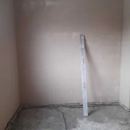 Pokój w trakcie prac wykończeniowych: częściowo otynkowana ściana, surowy beton na podłodze z widocznymi przewodami elektrycznymi przy ścianie, aluminiowa poziomica oparta o ścianę.
