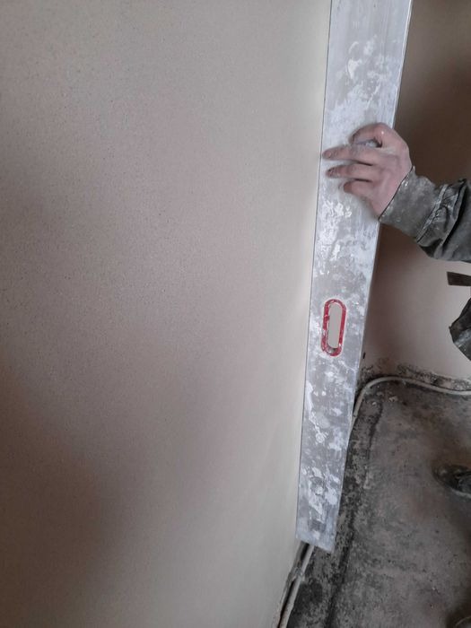 Ręka pracownika wygładza świeżo nałożony tynk maszynowy przy użyciu długiej, aluminiowej łaty; widoczna nierówna powierzchnia podłogi i listwa przypodłogowa.