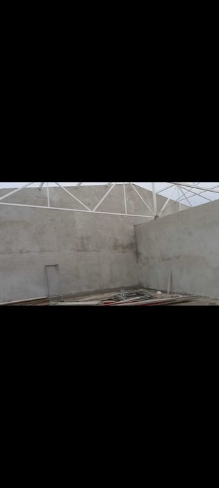 Wnętrze budynku w trakcie prac wykończeniowych: surowe, szare ściany po nałożeniu tynku, widoczna konstrukcja dachu i porozrzucane materiały budowlane w rogu.