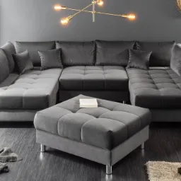 Szary, modułowy narożnik z pufą na tle ciemnej ściany, oświetlony lampą w stylu industrialnym, z dekoracjami na podłodze i ścianie.