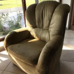 Oliwkowy fotel z wzorzystą tapicerką stoi na kafelkach w słonecznym pokoju z widokiem na ogród.