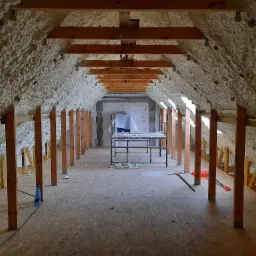 Poddasze ocieplone pianką PUR, widoczne drewniane belki konstrukcyjne i rusztowanie w tle, częściowo zakryte folią ochronną.