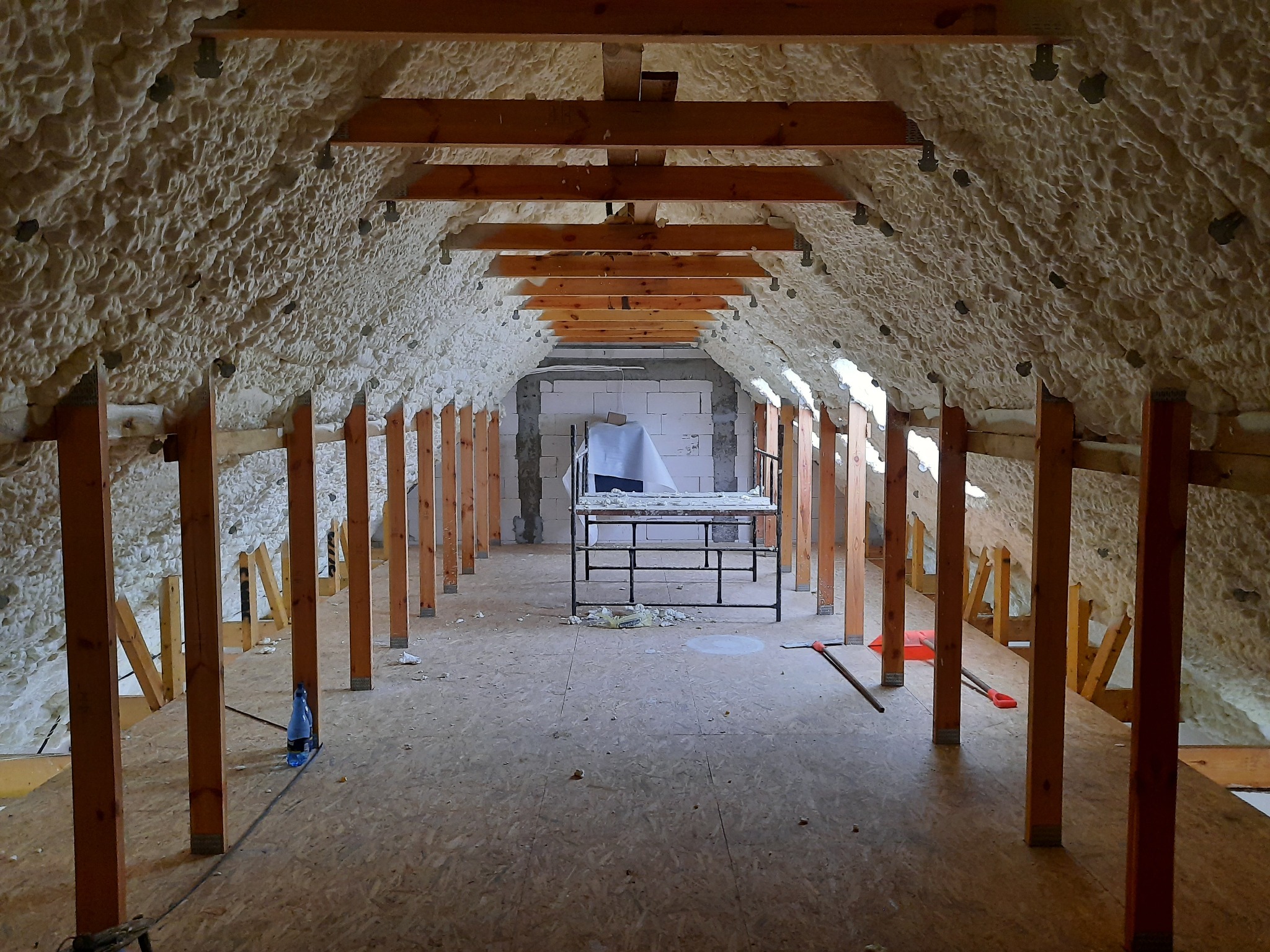 Poddasze ocieplone pianką PUR, widoczne drewniane belki konstrukcyjne i rusztowanie w tle, częściowo zakryte folią ochronną.