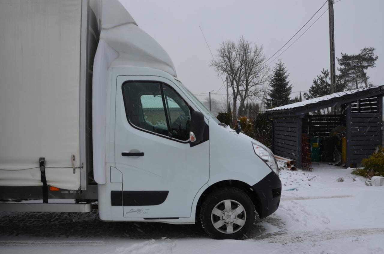 Biały samochód dostawczy typu bus z plandeką na tle zimowego krajobrazu z zaśnieżonym podjazdem i drewnianą wiatą.