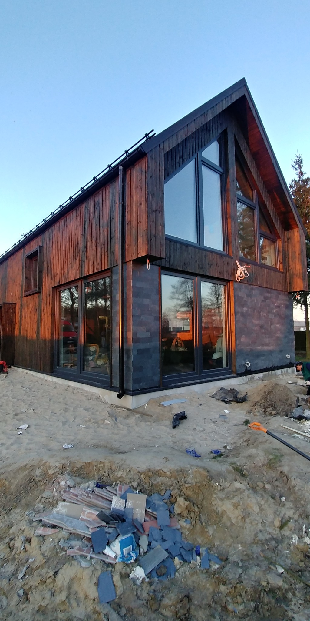 Nowoczesny dom z drewnianą elewacją w trakcie budowy, widoczne duże okna, ocieplenie fundamentów i pozostałości materiałów budowlanych na piaszczystym terenie.