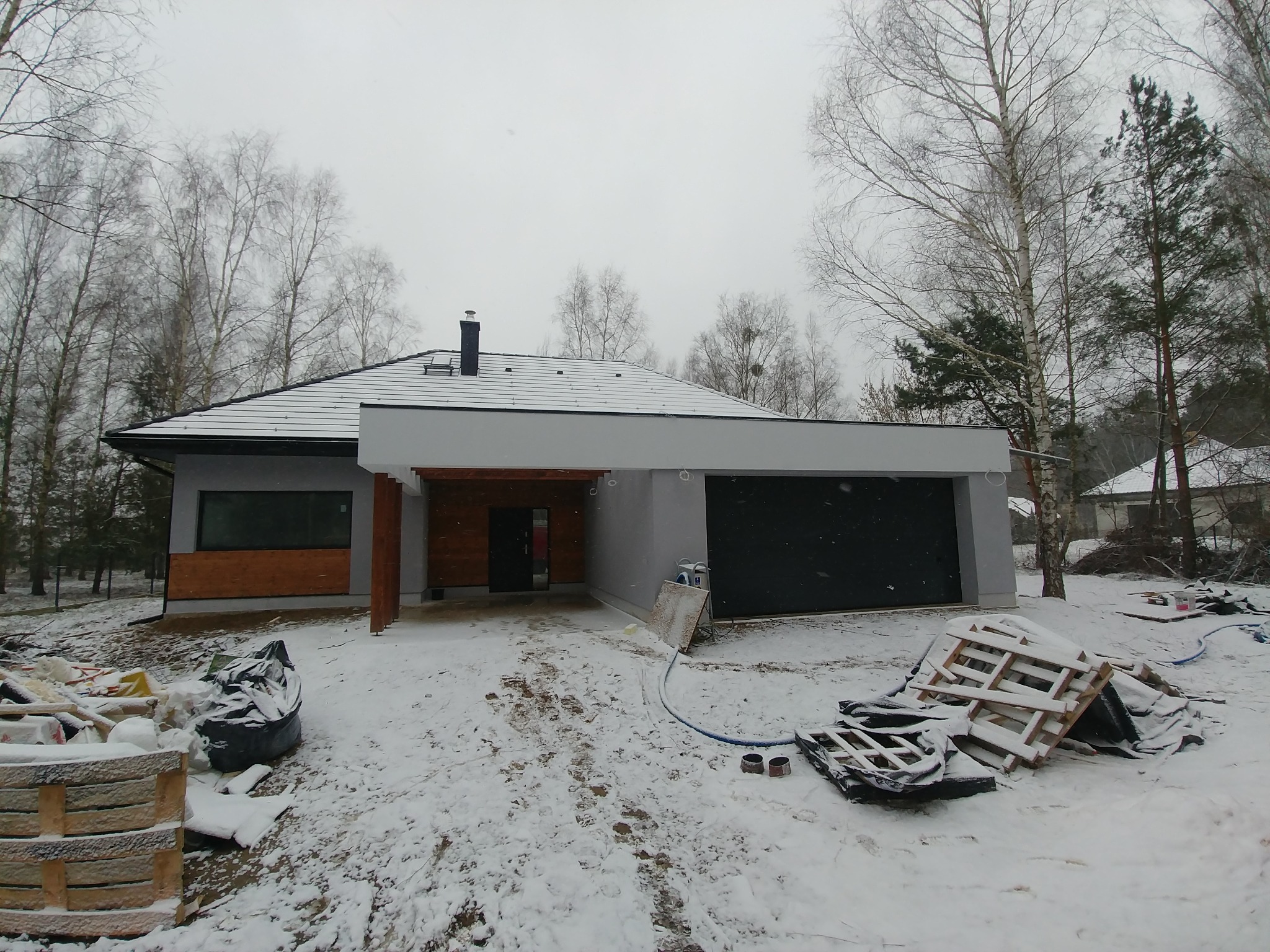 Nowoczesny dom z garażem w trakcie budowy, widoczne materiały budowlane na śniegu, drzewa bez liści w tle.