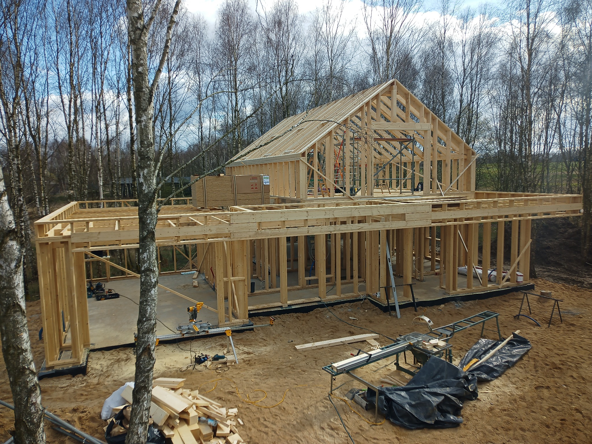 Wznoszenie drewnianej konstrukcji domu w stanie surowym otwartym, z widocznymi narzędziami i materiałami budowlanymi na placu budowy, w tle drzewa bez liści.