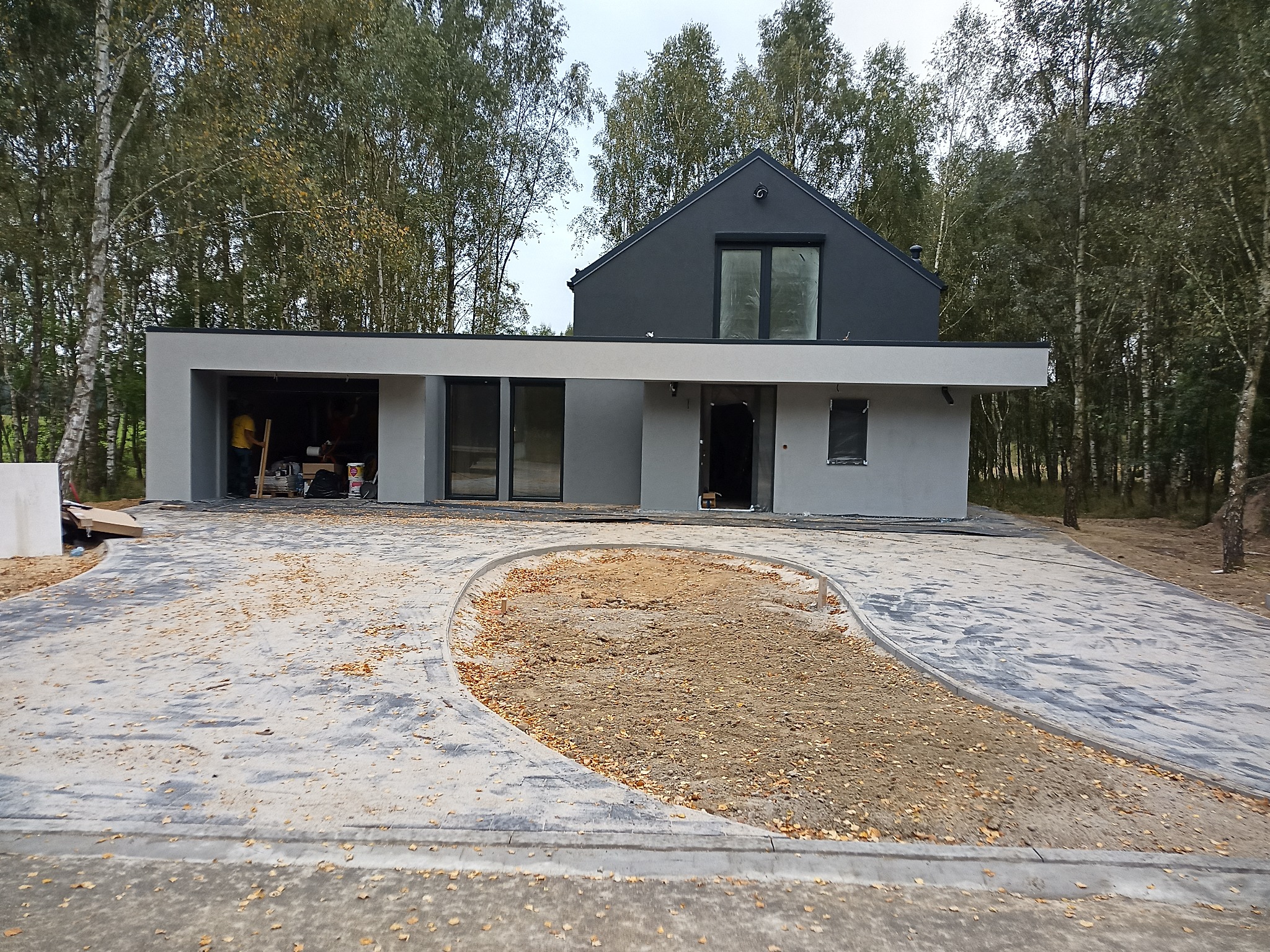 Nowoczesny dom z szarą elewacją w trakcie budowy, widoczny częściowo ułożony podjazd z kostki brukowej i przygotowany teren pod trawnik, prace wykończeniowe w garażu.